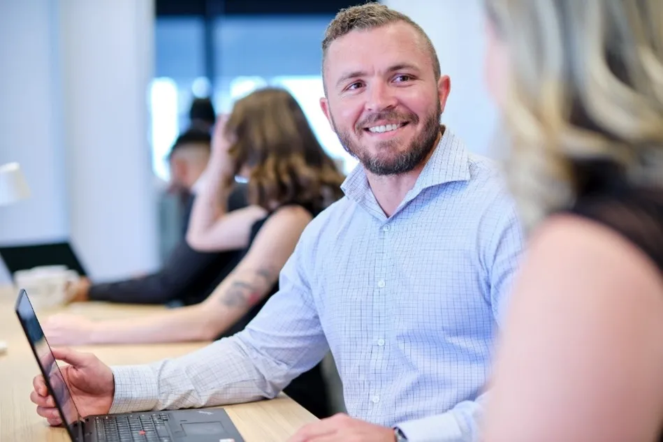 Male office worker in blue shirt with a beard looking up from computer and smiling