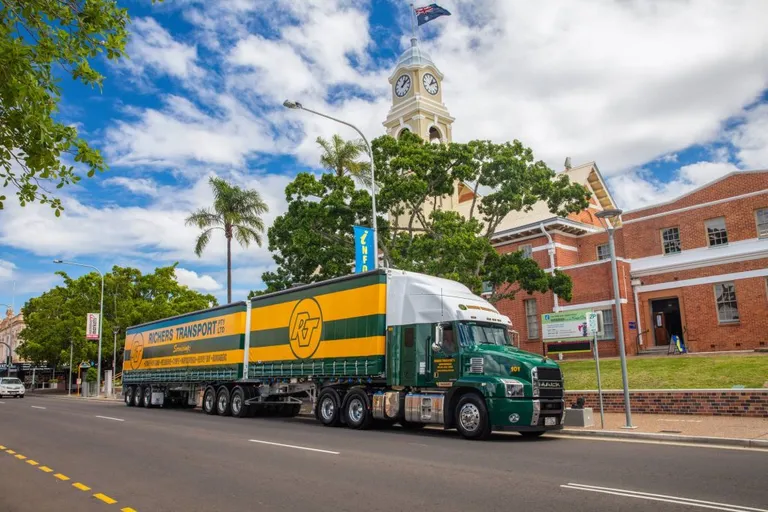 A Richers Transport B-double truck parked outside a heritage building. The truck has green and yellow stripes on the side, with the Richers Transport logo and branding.