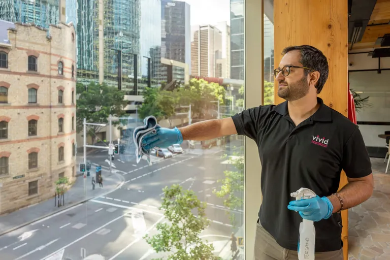 A male Vivid Services worker in a black t-shirt, cleaning an office window with a view out to a city street